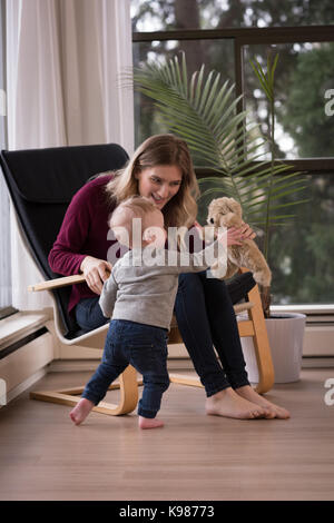 Glückliche Mutter, Stofftier zum Sohn beim Sitzen auf Stuhl von Fenster Stockfoto