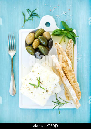 Frischem Schafskäse mit Oliven, Basilikum, Rosmarin und Scheiben Brot auf weißer Keramik mit Platine über hell türkis blau lackierten Holz- Hintergrund Stockfoto