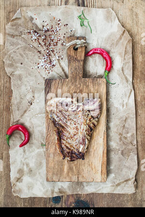 Gekochtes Fleisch T-Bone Steak auf dem Board mit roten Chilischoten, Gewürzen, frischem Rosmarin über ölige Handwerk Papier und rustikalen Holzmöbeln Hintergrund, selektiver Fokus Stockfoto