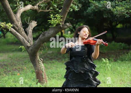 Eine schöne indische Mädchen Musiker in schwarzen Kleid spielen eine rote Violine vor einem Baum und Natur Stockfoto