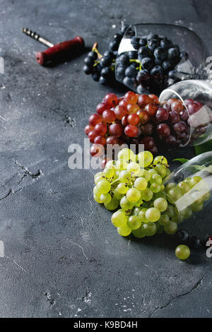 Vielzahl von drei Art frische reife Trauben dunkel Blau, Rot und Grün in verschiedenen Lügen Weingläser mit alten Korkenzieher über schwarzen Textur Hintergrund mit Stockfoto