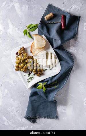 Weiße Platte mit in Scheiben geschnittenen Camembert Käse überbacken Bündel grüne Trauben serviert mit Brot, Wein Glas, Korkenzieher, grüne Blätter, Gabel auf Blau Textil Stockfoto