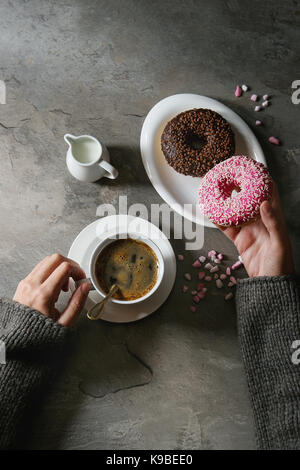 Platte von Bunt glasierter Krapfen mit rosa Zucker, Tasse schwarzen Kaffee, Kanne Milch über grau Textur Tabelle. weibliche Hände Donat. Flach mit sp Stockfoto