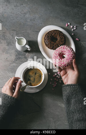 Platte von Bunt glasierter Krapfen mit rosa Zucker, Tasse schwarzen Kaffee, Kanne Milch über grau Textur Tabelle. weibliche Hände Donat. Flach mit sp Stockfoto