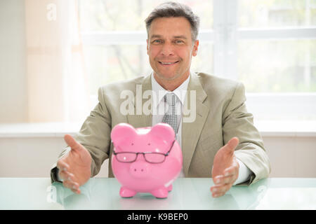 Portrait Of Happy Geschäftsmann mit Sparschwein Brillen tragen am Schreibtisch Stockfoto