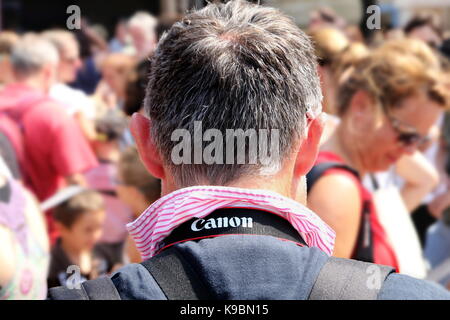 Zurück von einem mittleren Alter Mann, der eine Qualität gestreiften Hemd, und eine Canon Kamera um den Hals, vor einer Masse von Menschen in den unscharfen zurück Stockfoto