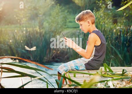 Sommer auf dem Land. Junge sitzt auf dem Steg über den See. Kleinen Fotografen unter Foto der Libelle von Smart Kamera des Telefons. Stockfoto
