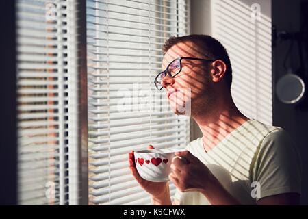 Nachdenklich (In der Liebe) Mann mit Brille schauen durch eine Jalousie beim Frühstück. Stockfoto