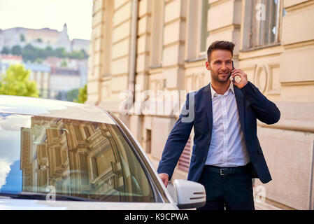 Ein hübscher junger Geschäftsmann zu sein Auto auf der Straße, während im Gespräch auf seinem Mobiltelefon Stockfoto