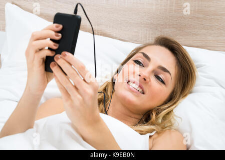 Glückliche junge Frau sitzt auf dem Bett Musik hören Stockfoto