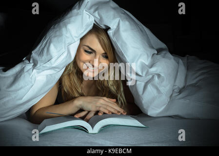 Glückliche junge Frau unter Betten lesen Buch Stockfoto