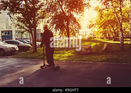 Alte grauhaarige Großvater, reiten Ihr scooter im Park in der Sonne bei Sonnenuntergang. Sport für Senioren. Skaten im Herbst in einem Stadtpark amo Stockfoto