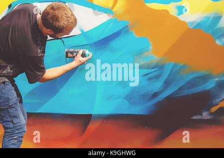 Foto in den Prozess der Ausarbeitung einer Graffiti Muster auf einem alten Betonwand. Die jungen Langhaarigen blonden Kerl zieht eine abstrakte Zeichnung in verschiedenen Farben. Stockfoto