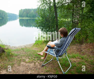 Junge sitzt im Liegestuhl ein Buch lesen auf einem See Stockfoto