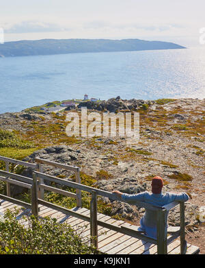 Mittlerer Erwachsener männlicher Tourist, der auf einem Holzsitz über dem Fishing Point (Fox Point) Lighthouse, St. Anthony, Great Northern Peninsula, Neufundland, Kanada sitzt Stockfoto