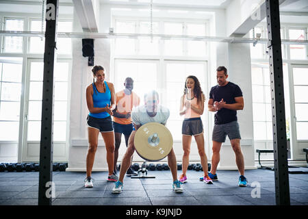 Lächelnden jungen Mann belasten Gewichte in einem Fitnessraum mit einer vielfältigen Gruppe von Freunde ihn Anfeuern im Hintergrund zu heben Stockfoto
