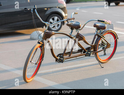 Eine Schokolade - farbige tandem Fahrrad mit Scarlet Räder ist in einem Parkhaus in der Nähe der Radweg in der Abendsonne geparkt. Stockfoto