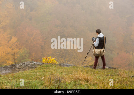 Natur, Arbeit, Hobby Konzept. junger Mann in warme Kleidung angezogen, Burgund Hose, Grau Sweatshirt und schwarze Weste, neben mit Kamera auf Stativ und die Bilder von Herbst Wald Stockfoto
