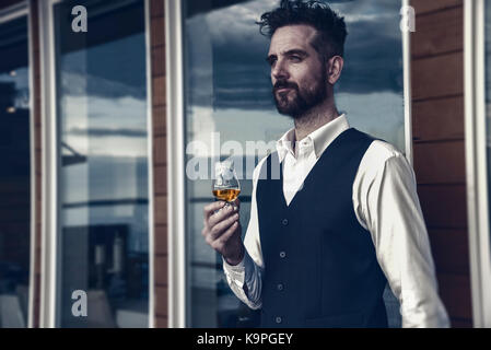 Sehr schön im mittleren Alter kaukasischer Mann mit Bart Holding altmodische Whiskey Glas blickt auf eine ominöse Sky von Deck mit Glasfenstern. Stockfoto