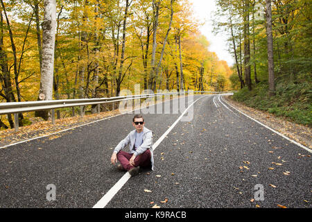 Reisen, Meditation, Freiheit Konzept. Modisch und friedlicher Mann in der Lotus Position sitzt rechts auf ununterbrochenen Linie von der Straße, die durch den Wald Stockfoto