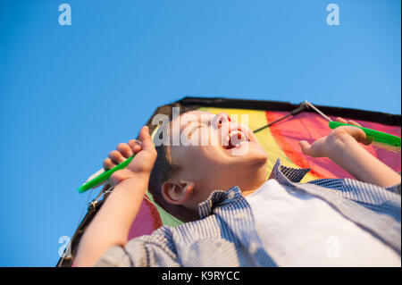 Lachender fröhlicher Junge, der einen Drachen über den Kopf gegen einen blauen Himmel hält Stockfoto