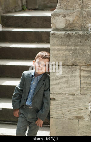 Schöner kleiner Junge in einem Kostüm, der sich an eine alte Steinmauer lehnt Stockfoto