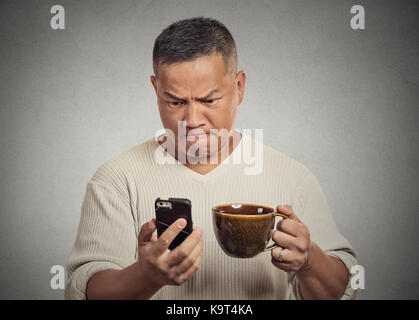 Closeup Portrait besorgt wütend frustrierter Mann lesen schlechte Nachrichten sms auf Smart Handy trinken Holding Tasse Kaffee Kaffee graue Wand Hintergrund isoliert Stockfoto