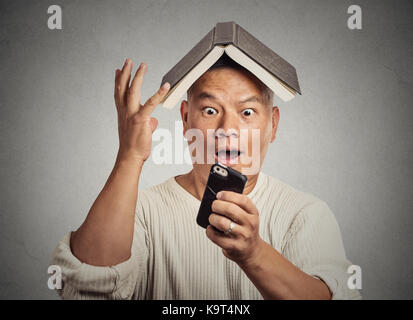 Porträt Porträt überrascht betäubt im mittleren Alter Mann komisch aussehende Kerl mit Buch auf dem Kopf news Lesen auf Smart Phone isoliert grauen Hintergrund. Hum Stockfoto
