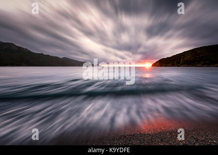 Slow Shutter Bild der Wellen auf einem Kieselstrand, wie die Sonne am Bussaglia an der Westküste von Korsika unter dunklen Gewitterwolken Stockfoto