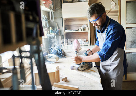 Portrait von Reife bärtige Tischler polieren Stück Holz in der Werkstatt gegen Fenster und Möbel Stockfoto