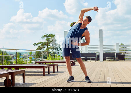 Aktive junge Mann in Sportswear genießen Sie den atemberaubenden Blick, während stretching Arme vor Outdoor Training im geräumigen Terrasse Stockfoto