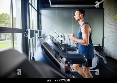 Profil anzeigen von Konzentriertem passen Mann Hören von Musik über Kopfhörer während der Ausführung auf Laufband im modernen Fitnessraum mit Panoramafenstern, Portraitfotos Stockfoto