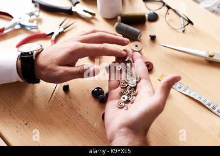 Nahaufnahme der männlichen Tailor holding Snap Buttons in der Hand, arbeiten Holztisch mit Schere, Maßband, Gewinde Spulen und andere Nähen Elemente auf Stockfoto