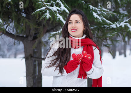 Mädchen im Winter Wald mit roten Herzen in die Hände. Winterurlaub, Valentinstag, Personen Konzept. Winter outdoor Porträt der jungen Frau in Pullover Stockfoto