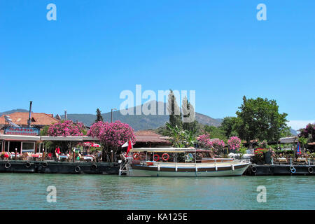 Urlauber in einem mit Oleander-Blumen bedeckten Restaurant neben dem Ausflugsboot entlang der hübschen Uferstadt Dalyan, Türkei Stockfoto