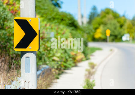 Schwarz auf Gelb chevron Schild angebracht zu posten, zeigt rechts gegen blurry geschwungene Straße im Hintergrund. Stockfoto