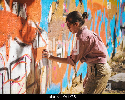 Schönen jungen Mädchen einen bunten Graffiti mit Sprühdose auf Urban Street Wall. Filmische getönten erschossen. Kreative Art. begabte Frau in Gläsern Stockfoto