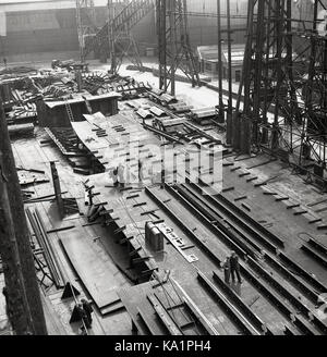 Anfang der 1950er Jahre, historisches Bild von J Allan Cash, das Arbeiter am Boden zeigt, die von den riesigen Gantries der Werft in Harland & Wolff in Belfast Docks, Nordirland, in den Schatten gestellt wurden, die für den Standort berühmt waren, an dem der Titantic-Ozeandampfer gebaut wurde. Stockfoto