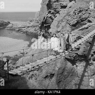 Ein Mann und zwei Damen aus den 1950er Jahren, die auf der Carrick-a-Rede-Seilbrücke, einer berühmten Seil- oder Schaukelbrücke in der Nähe von Ballintoy in der Grafschaft Antrim, Nordirland, stehen, die erstmals 1755 vom lokalen Lachsfischer errichtet wurde. Stockfoto