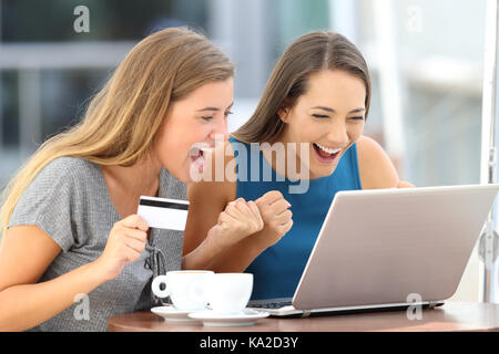 Zwei aufgeregt Freunde entdecken bietet Kaufen auf Linie mit einem Laptop und einer Kreditkarte in einer Bar auf der Terrasse sitzen Stockfoto