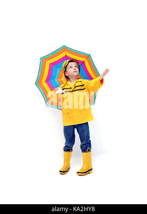 Junge in gelb Regenjacke und Gummistiefel hält seine Hand für Regen zu prüfen. Er hält ein bunt gestreiften Schirm. Stockfoto