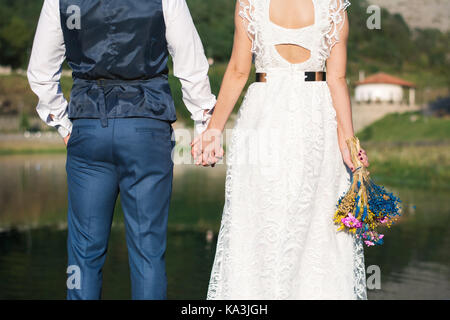 Braut und Bräutigam halten sich an den Händen vor einem See Stockfoto