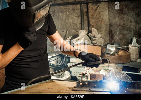 Ein erwachsener Mann Schweißer in einem schwarzen T-Shirt und eine schwarze Maske brüht einen Metall schweißen Schweißgerät in einem dunklen Werkstatt Stockfoto