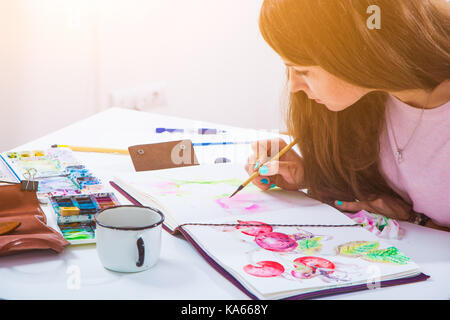 Dunkelhaarige Künstlerin mit hellen Nägel zeichnet eine dünne Holz- Pinsel und Aquarellfarben im Album zum Zeichnen auf weißem Papier rosa Pfingstrosen, auf dem Tisch liegen w Stockfoto