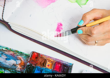Die Nahaufnahme als Künstler mit blauen Nägel zieht rosa Blüten mit einem hölzernen Pinsel und Aquarellfarben in der aluoma für Zeichnung, auf dem Tisch liegen eine Palette f Stockfoto