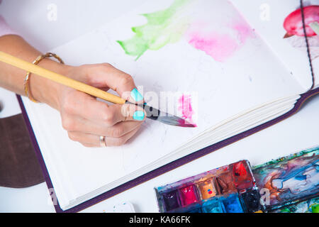 Die Nahaufnahme als Künstler mit blauen Nägel zieht rosa Blüten mit einem hölzernen Pinsel und Aquarellfarben in der aluoma für Zeichnung, auf dem Tisch liegen eine Palette f Stockfoto