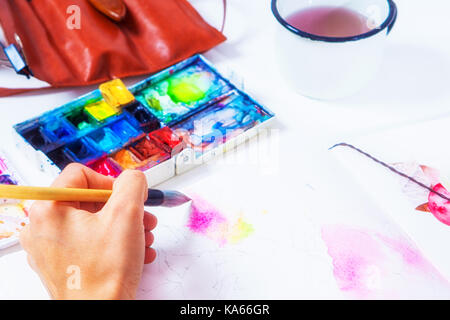 Ein Close-up Künstler malt rosa Blüten mit einem hölzernen Pinsel und Aquarellfarben in der Zeichnung Album, auf dem Tisch liegen eine Zeichnung Palette, eine Ledertasche mit Stockfoto