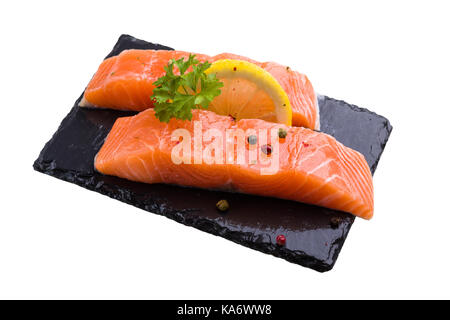 Zwei Stücke von Lachs roher Fisch, dekoriert mit Zitronenscheibe, Petersilie und Pfeffer würzen, auf schwarzem Schiefer legen, auf weissem isoliert. Stockfoto