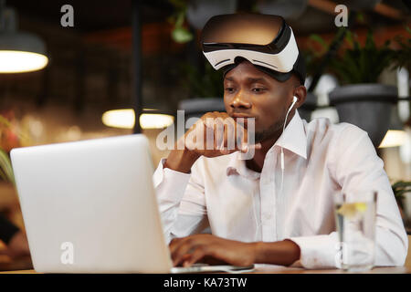 Ernsthafte afrikanischer Mann ich Virtual Head Set, mit Laptop. Stockfoto