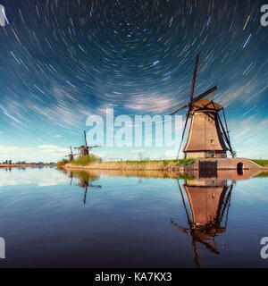 Bunte Feder Nacht mit traditionellen niederländischen Windmühlen Kanal in Rotterdam. Hölzernen Pier in der Nähe der See. Holland. Niederlande. Fantastischen Sternenhimmel und die Milchstraße. Stockfoto